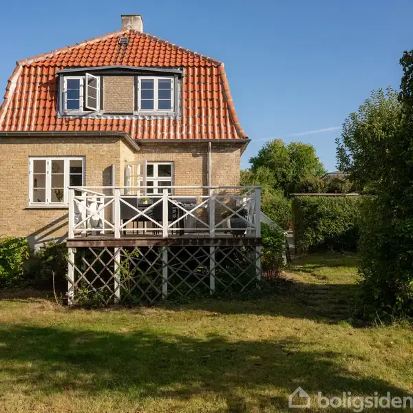 Et murstenshus med rødt tegltag har en hvid træterrasse på forsiden. Husets vinduer er åbne, og det er omgivet af en grøn have under en klar, blå himmel.