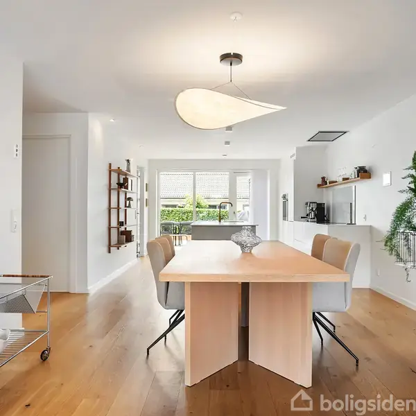 Et lyst spisestueområde med et moderne bord omgivet af fire stole. En dekorativ lampe hænger over bordet. Rummet er omgivet af en minimal indretning med planter og hylder.
