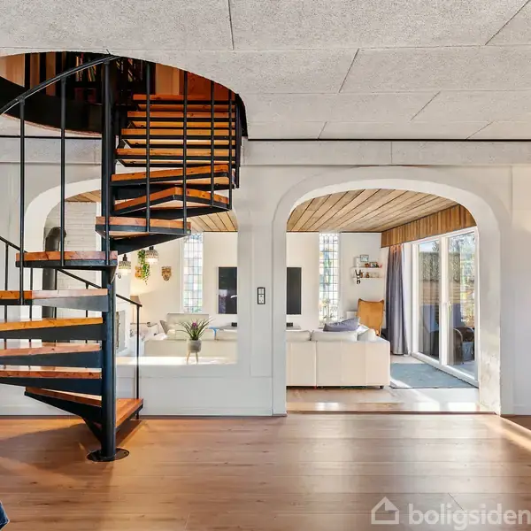 En vindeltrappe i metal og træ fører op til næste etage. Den står i en stue med trægulv, buede døråbninger og moderne indretning, omkranset af bøger og sofaer.