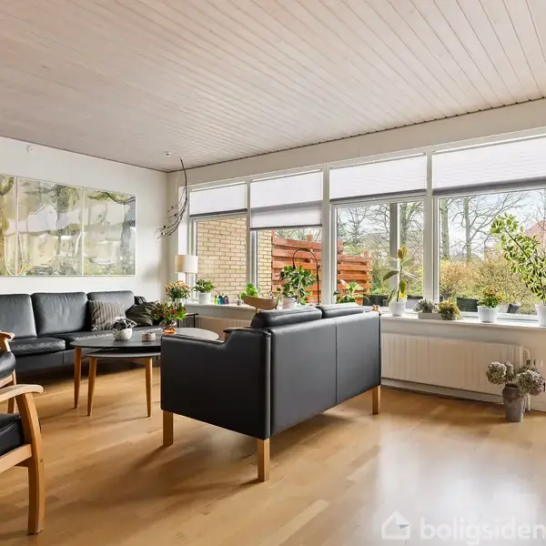 Stue med sorte sofaer og træstole står omkring et lavt bord. Store vinduer lader lys ind, og planter pynter vindueskarmen. Udendørs vises en have og terrasse.