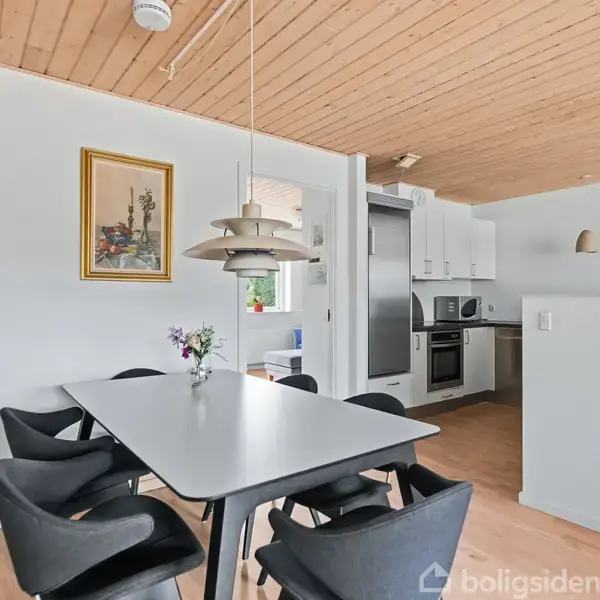 Spisebord med sorte stole omgivet af et lyst køkken. Bordet har en vase med blomster. En moderne lampe hænger over bordet. Maleri på væggen.