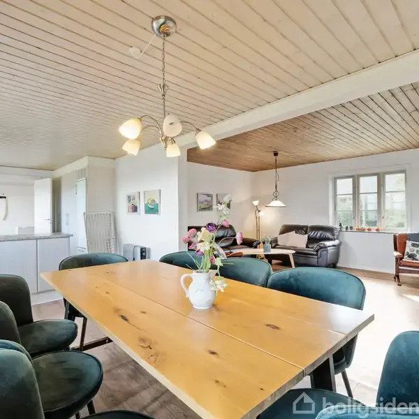 Et spisebord med en vase og blomster står i midten omgivet af grønne stole i en lys stue-køkken alrum. Der er hvidt køkken til venstre og sofagruppe til højre.