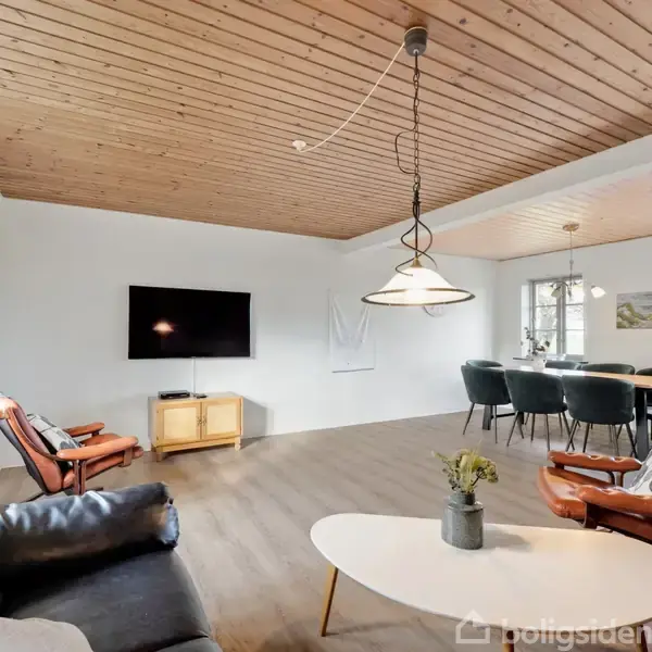 Stue med sort lædersofa, hvidt rundt bord og TV på væggen. Spisebord med seks stole i baggrunden. Træloft og trægulv skaber en lys og indbydende atmosfære.