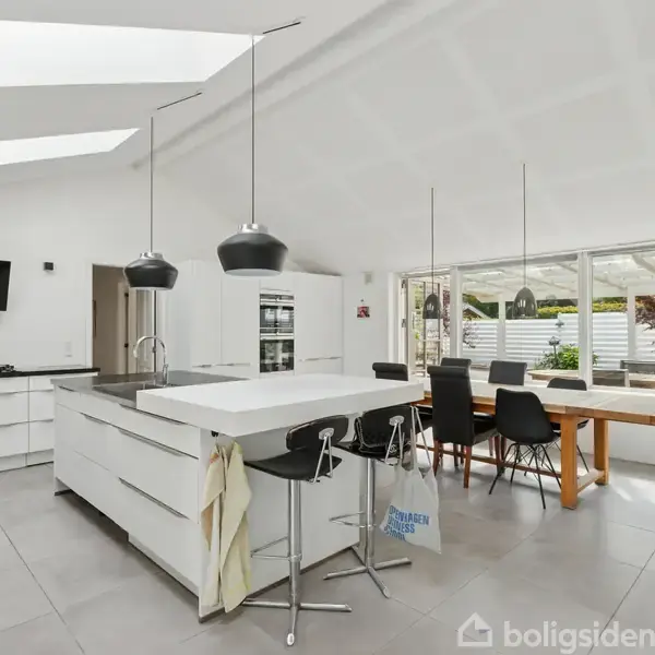 Køkken med hvid køkkenø og sort barstol står i moderne, lys spisestue med store vinduer og spisebord med sorte stole under hængende lamper. Tasker hænger på køkkenøen.