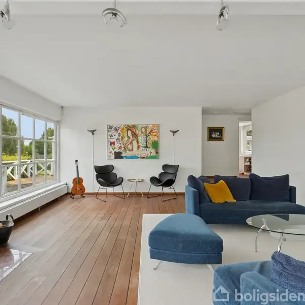 Brændeovn opvarmer moderne stue med trægulv og blå sofaer. Store vinduer giver udsigt til grøn have, mens en guitar står ved væggen. Væg malet med farverigt billede.