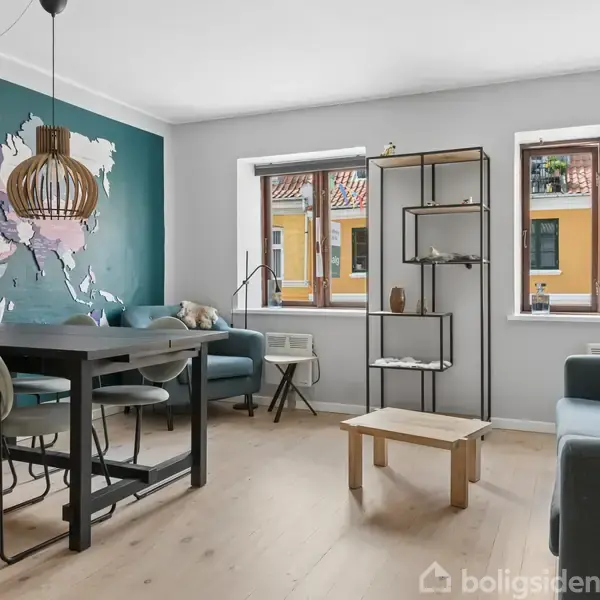 En stue med spisebord har en verdenskort-væg og pendellampe. To vinduer giver udsigt til nabohuse. Sofa, sofabord, og reol fuldender rummet. Vinduerne slipper lyset ind.