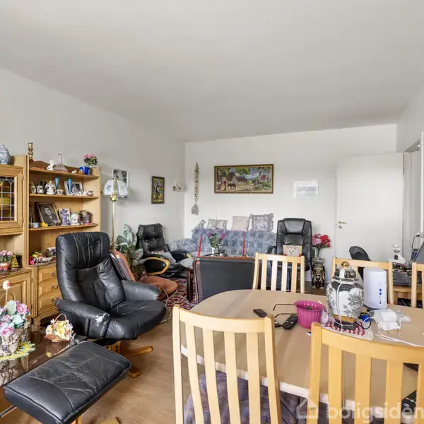 Stue med spisebord omgivet af træstole, lænestole og sofa. Skænk med dekorationer står langs væggen. Rummet er lyst med hvide vægge og dekoreret med billeder.