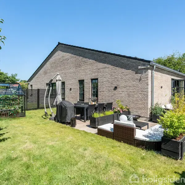 Et murstenshus med en terrasse, der har havemøbler og en grill, placeret i en grøn have med planter i krukker og et drivhus i baggrunden.