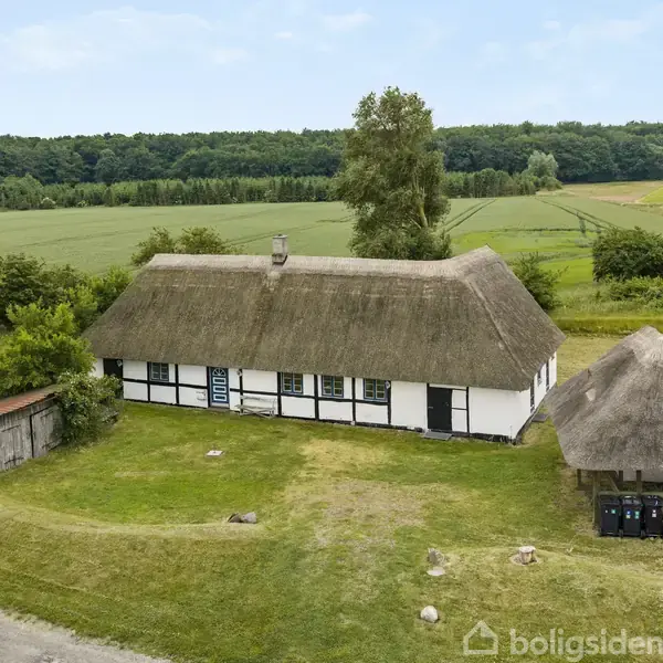 Bindingsværkshus ligger stille på en græsmark omgivet af skov og marker. Ved siden af huset står en stråtækt pavillon og en lille træskur. Ingen tekst fundet i billedet.