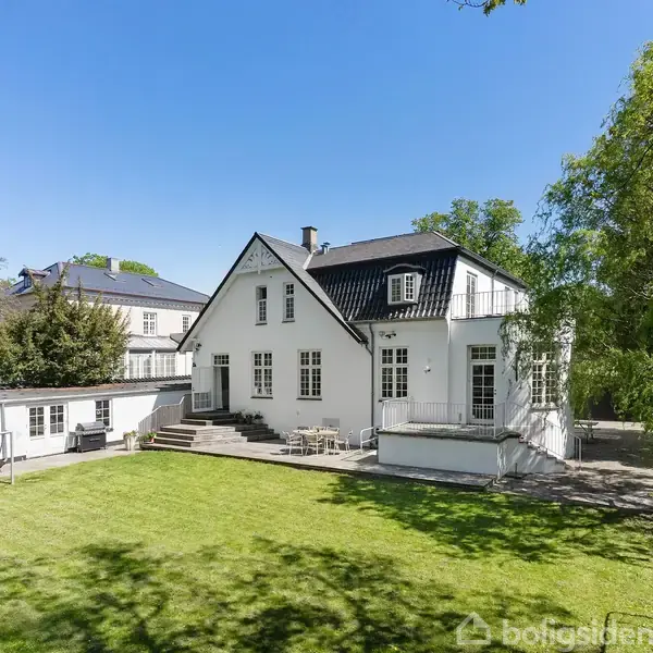 Hvidt hus med sort tag står i solrigt vejr omgivet af grøn græsplæne og træer. Terrace med havemøbler foran huset indikerer afslapning i en velplejet have.