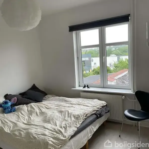 Seng med puder og en blå bamse ligger i et værelse. Ved siden af er et vindue med udsigt til huse og træer. En stol står ved kanten af billedet.