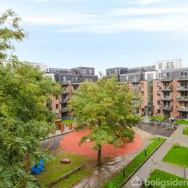 Legeplads med rutsjebane står stille omgivet af boligkomplekser. En stor træ står midt på en rund, rød flade. Græsplæner og træer danner en grøn ramme.