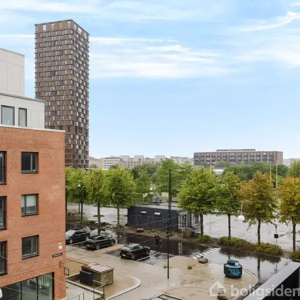 Bygning står stille blandt andre strukturer, omgivet af parkerede biler og træer, med en høj bygning i baggrunden. Skyfri blå himmel.