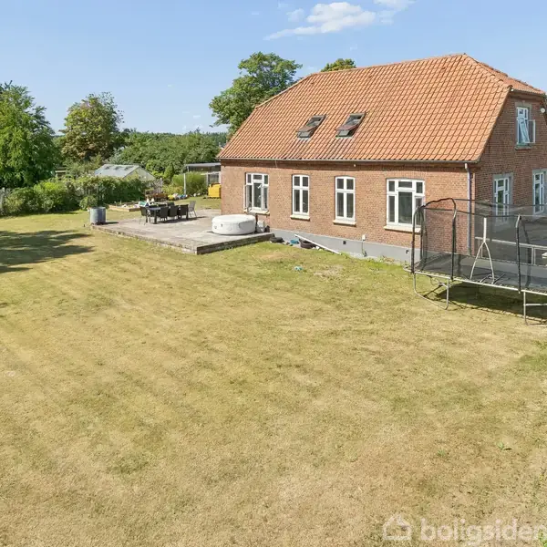 Murstenshus med rødt tegltag står i en stor, velplejet græsplæne. En trampolin er placeret i haven ved siden af en terrasse med havemøbler. Solrigt vejr med blå himmel.