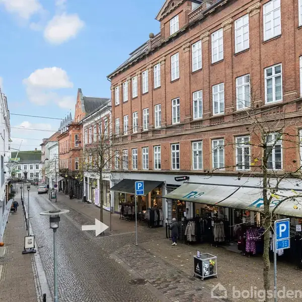 En bred gågade med brosten viser butikker med udstillingsvinduer og tøjstativer langs fortovet. Røde murstensbygninger med hvide vinduer omgiver gaden under en blå himmel med få skyer.