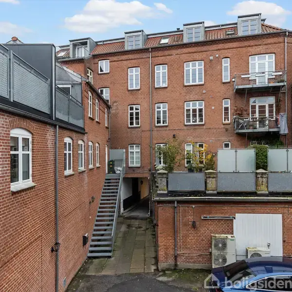 Rød murstensbygning, med flere vinduer og altaner, står i en gård omkranset af andre bygninger. En metaltrappe fører til en dør, og køretøj ses delvis i forgrunden.