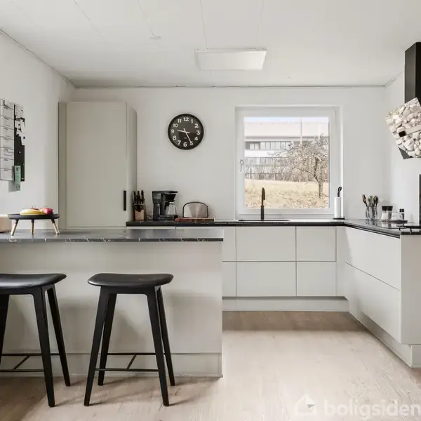 To moderne barstole står ved en køkkenø i et lyst køkken med hvidlige skabe. En kaffemaskine, knivblok, og ur pryder bordpladen. Store vinduer giver udsigt til en have.