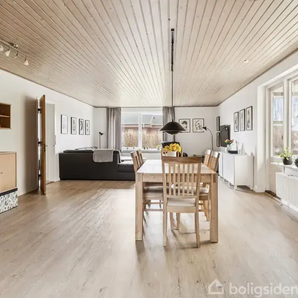 Spisebord med seks stole står midt i en lys stue. Rummet har trægulv og træloft med sorte lamper over bordet. Vinduerne til højre slipper naturligt lys ind.