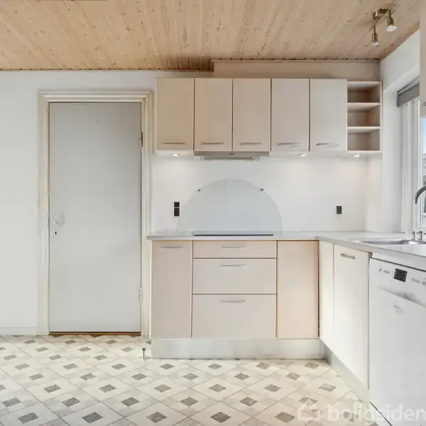 Køkkenskabe hænger over en vask ved en hvid bordplade. En ovn er indbygget i væggen til venstre. Gulvet har et ternet mønster, og et vindue lyser rummet op.