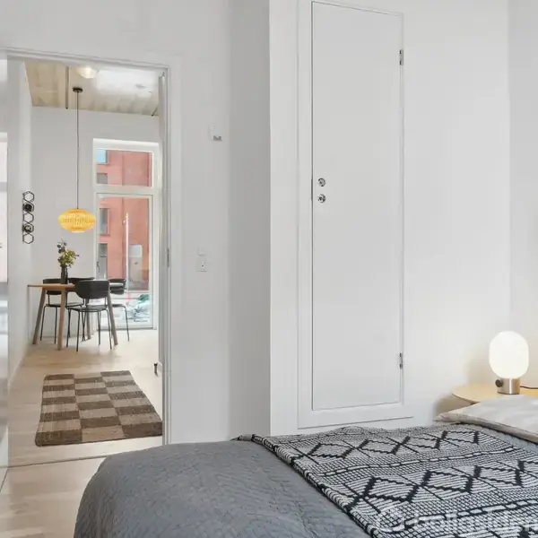 Soveværelse med en redt seng, en lampet på natbordet ved siden af. I baggrunden ses en åben dør, der fører til en spisestue med et bord, stole og hængende lampe.