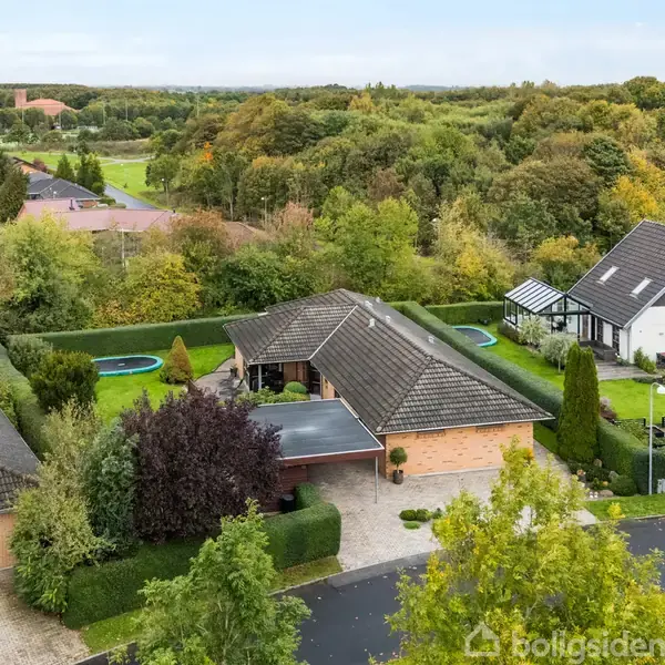 Et enfamiliehus med fladt tag står omgivet af grønne områder og haver i et roligt villakvarter. Nabohuse og skovkant udgør den omkringliggende natur.