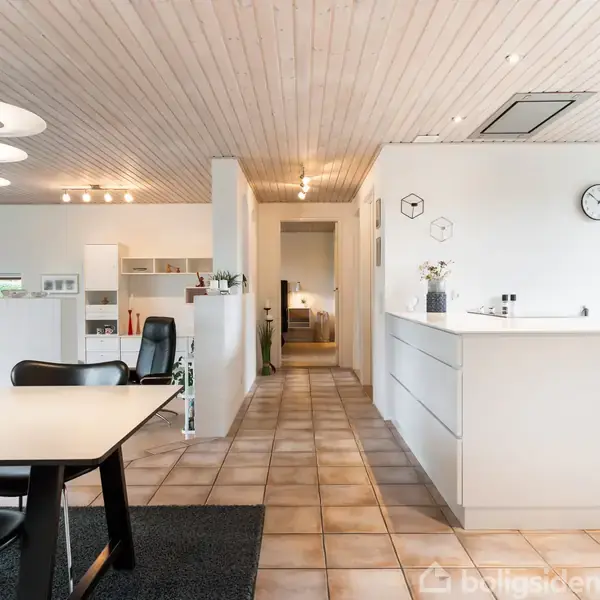 Et moderne køkken med hvidt interiør og sorte stole omkring et bord. Flisegulve og træloft udgør rummet, der er lyst op af pendellamper og spotlys, samt en vægmonteret ur.