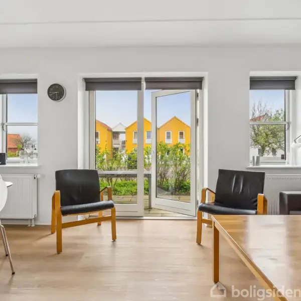 Stue med to stole og et sofabord vendt mod dobbeltdøre, der åbner ud til en have. Der er også et spisebord med stole og udsigt til gule huse udenfor.