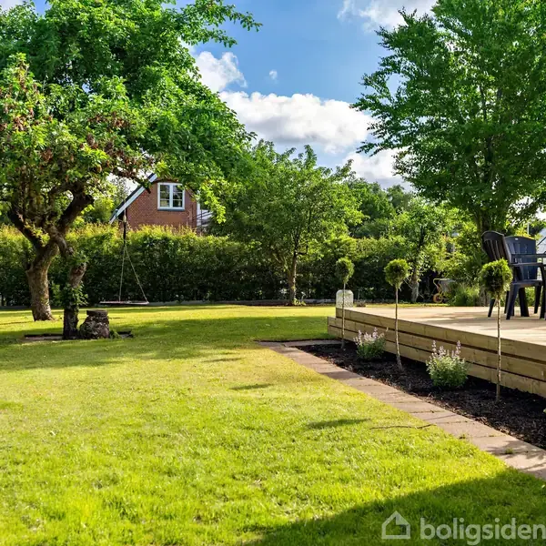 Græsplæne med terrasse og havemøbler, omkranset af træer og buske. Et rødt hus er synligt i baggrunden under en blå himmel med skyer.