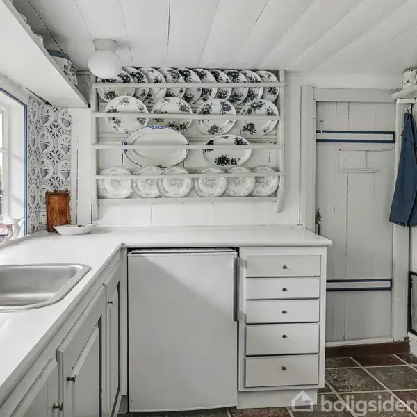 Hvidt køkken med blåmønstrede fliser. En stålvask til venstre, hylde med porcelænstallerkener på væggen. Lille køleskab og skuffer under bordplade. Dør i baggrunden. Garderobe med ophængt jakke.