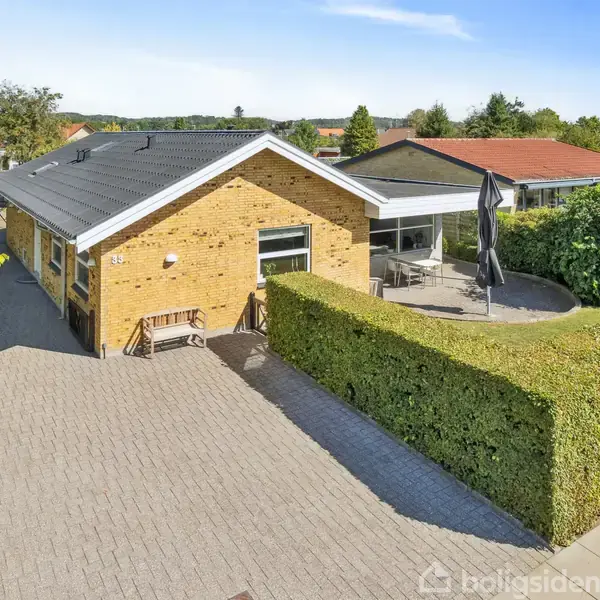 Et gult murstenshus med sort tag præsenterer en stor solbeskinnet terrasse omkranset af en trimmet hæk. Et bord med parasol står på terrassen i den rolige boligkvarter.