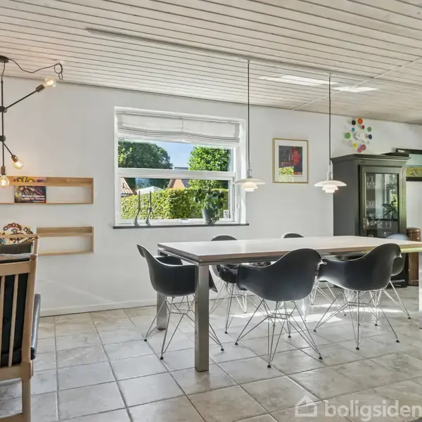 Spisebord står i et lyst rum med fliser på gulvet. Sorte stole omringer bordet. Store vinduer og vægdekorationer omgiver rummet, der har et moderne, minimalistisk præg.