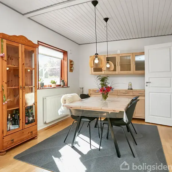 Et spisebord omgivet af moderne stole står på et tæppe i et lyst rum. En vitrineskab og en kommode med skuffer er i baggrunden ved et vindue.