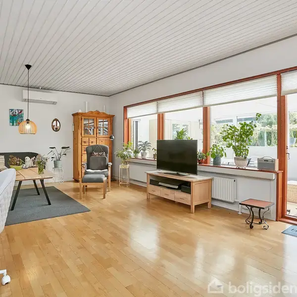 Stue med trægulv og TV på skænk. Aktuel handling: Sollys strømmer ind gennem store vinduer. Kontekst: Plantepynt, sofa, reol og spisebord skaber en hyggelig atmosfære.