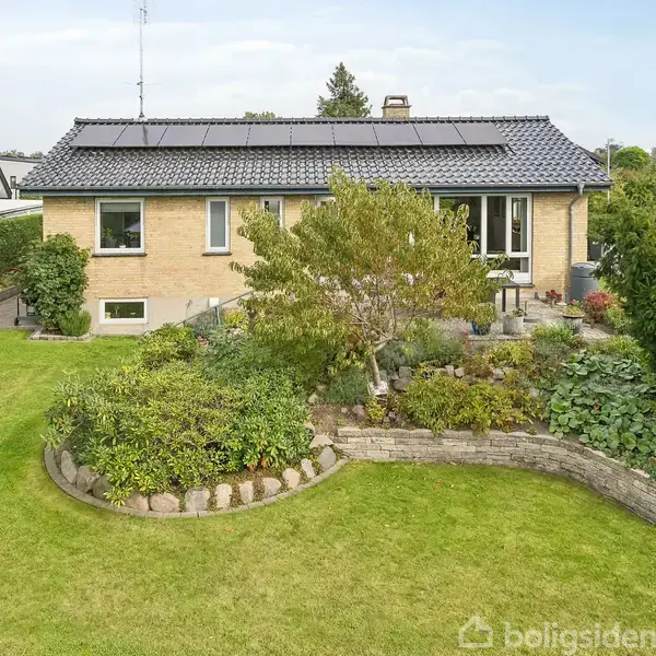 Hus med solpaneler på taget, omgivet af en velplejet have med græsplæne og blomsterbede. Ligger i et boligområde med huse i nærheden.