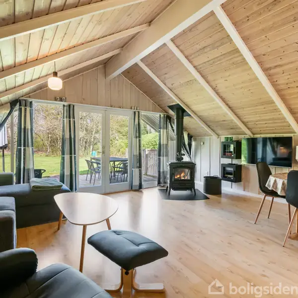 Stue med skråt loft og trægulv, indeholder sort sofa, brændeovn og spisebord. Store vinduer åbner ud mod en terrasse og have.