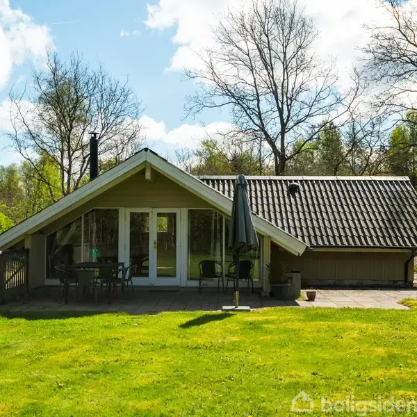 Sommerhus med skrå tag ligger i grøn have. Foran huset en terrassemøbleret med parasol. Omgivet af buske og træer under en klar blå himmel.