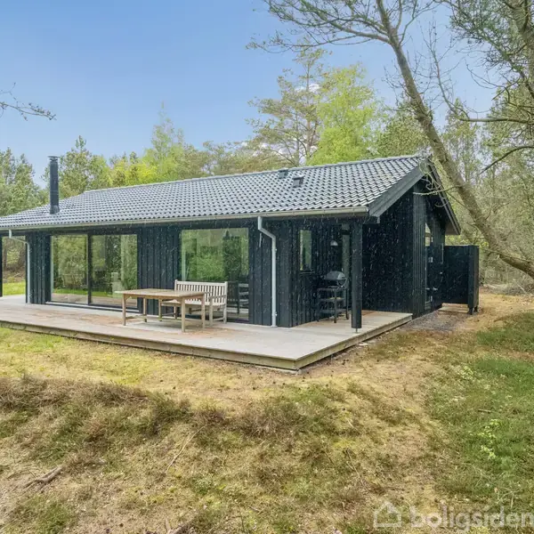 Sort træhus med terrasse. Et bord og bænke står udenfor. Omgivet af græs og træer i en skovrig område.