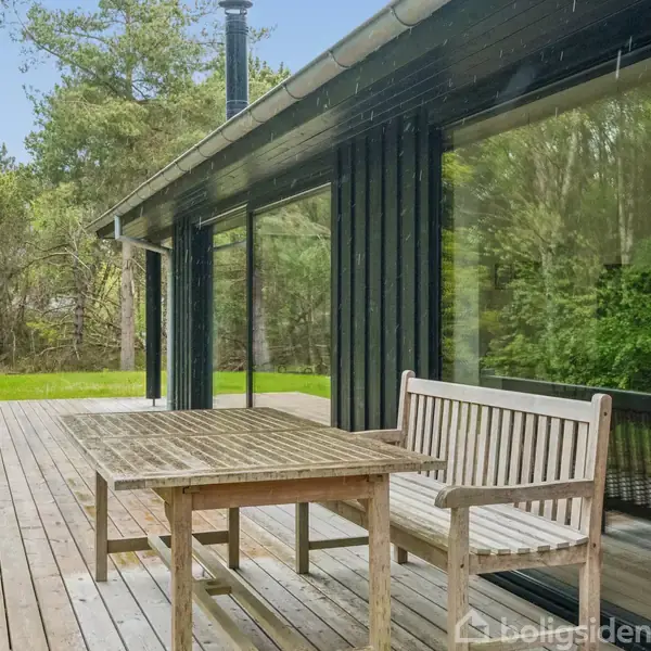 Træbord og bænk står på en træterrasse udenfor et sort hus. Store vinduer reflekterer skovens træer i baggrunden, hvilket skaber en rolig, naturlig atmosfære.