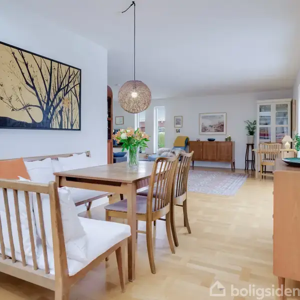 Spisestue med træbord og stole arrangeret. En blomsterbuket er centralt placeret. Væggen har et kunstværk, og der er et skab samt stole i baggrunden.