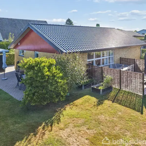 Et gult murstenshus med sort tag ligger i en grøn have. Foran huset er en terrasse med borde, stole og en parasol. Omkringliggende huse ses i baggrunden under en klar blå himmel.