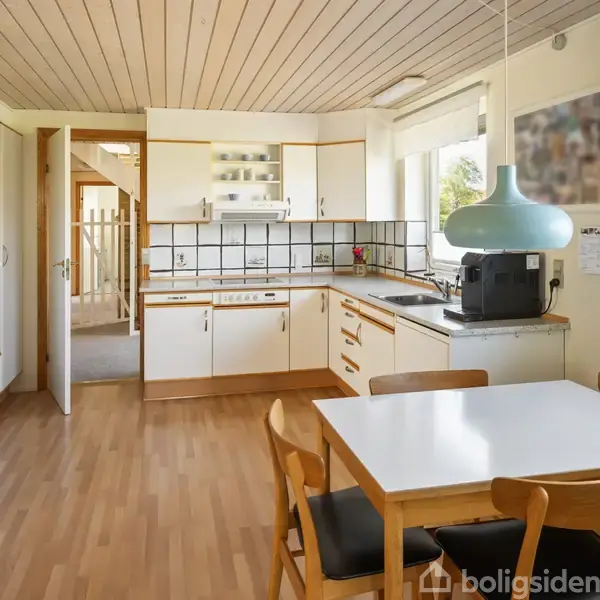 Køkken med hvide skabe og stålgreb, spisebord med fire stole i forgrunden, stående kaffemaskine og ovn. Træloft og vinduer giver lys, en tavle hænger på væggen.