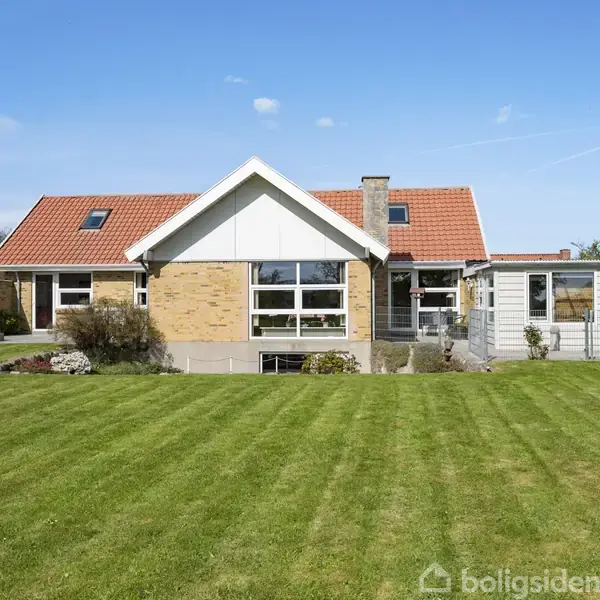 Et hus med rødt tag står i en velplejet have med grøn græsplæne. Huset har store vinduer og en tilstødende udestue. Det er omgivet af en blå himmel.