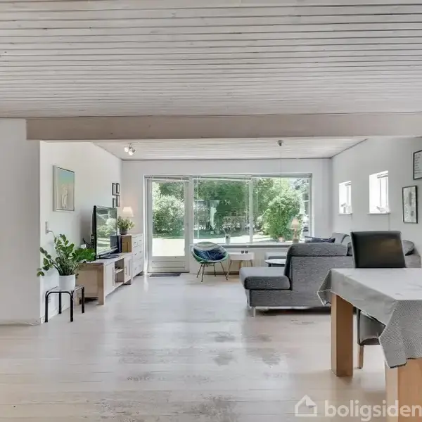 Stue med lyst trægulv og træloft; møbler inkluderer spisebord med sorte stole, grå sofa, og vitrineskab; store vinduer åbner ud mod en have.