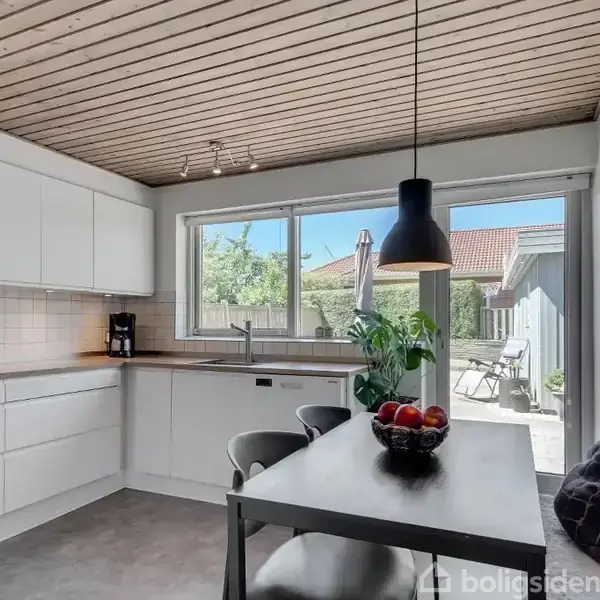 Et moderne køkken med hvide skabe har et spisebord med en frugtskål. Et stort vindue viser en terrasse med møbler udenfor. Det lyse rum har et træloft.