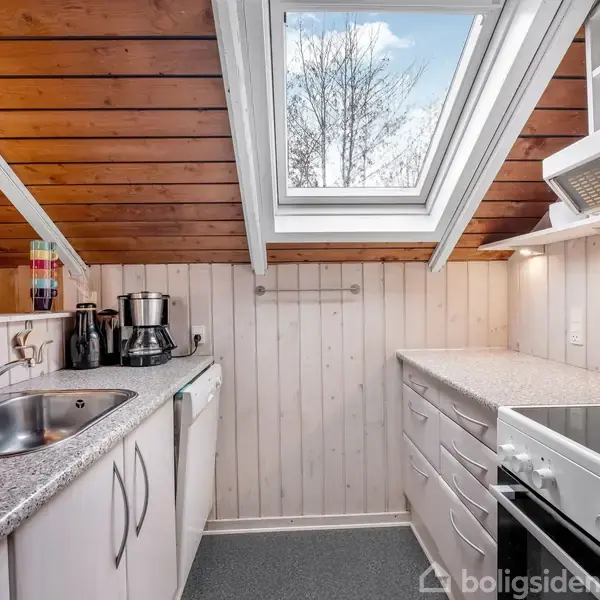 Et lille køkken med skråloft har hvidmalede trævægge, en vask og ovn på hver side, og loftsvindue der giver naturligt lys. Der er også køkkenredskaber på en bordplade.
