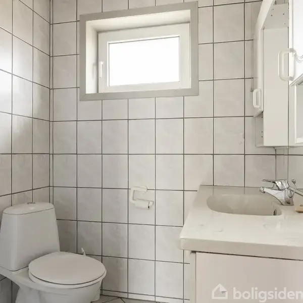 Toilet er placeret i et flisebelagt badeværelse ved siden af en vask med et spejlskab over. Et lille vindue giver lysindfald. Det ser rent og organiseret ud.
