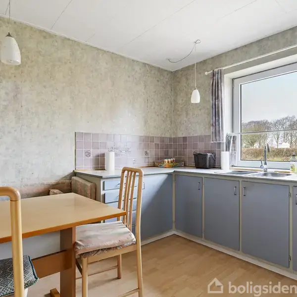 Et køkken med lyseblå skabe og beige bordplade. Et firkantet spisebord med træstole står i midten. Et stort vindue giver udsigt til træer udenfor.