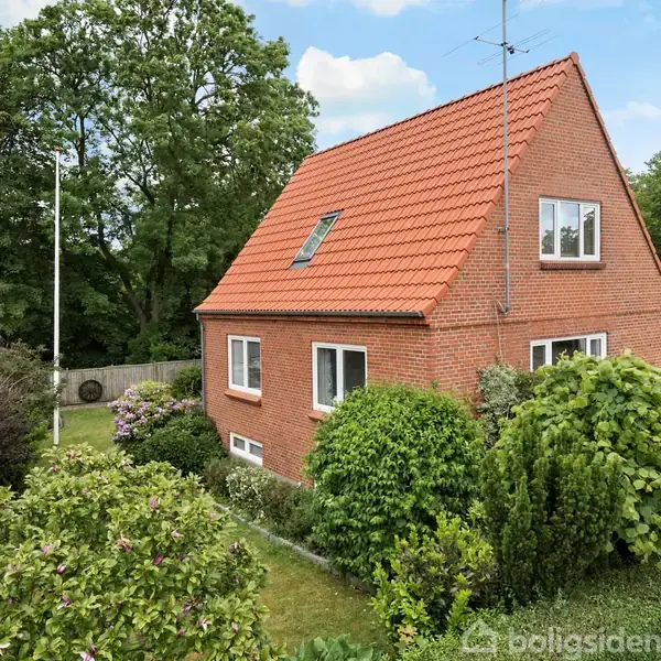 Rødt murstenshus står stille i en frodig have med flere buske og træer. Husets tag er orange, og der er en antenne på taget. Himlen er delvist skyet.
