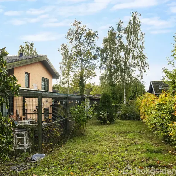 Hus med rød murstensfacade står stille i en have med græsplæne omgivet af hække og træer. Et nydeligt, åbent skyområde ses i baggrunden.