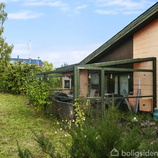 Et rødstenshus med skråt tag har en indhegnet terrasse. Omkringliggende have med grønne buske og træer. Himmel er blå med få skyer. Græsplænen er velplejet.